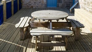 Un bâtiment en pierre avec une porte bleue, une terrasse en bois avec des tables de pique-nique et une cheminée.