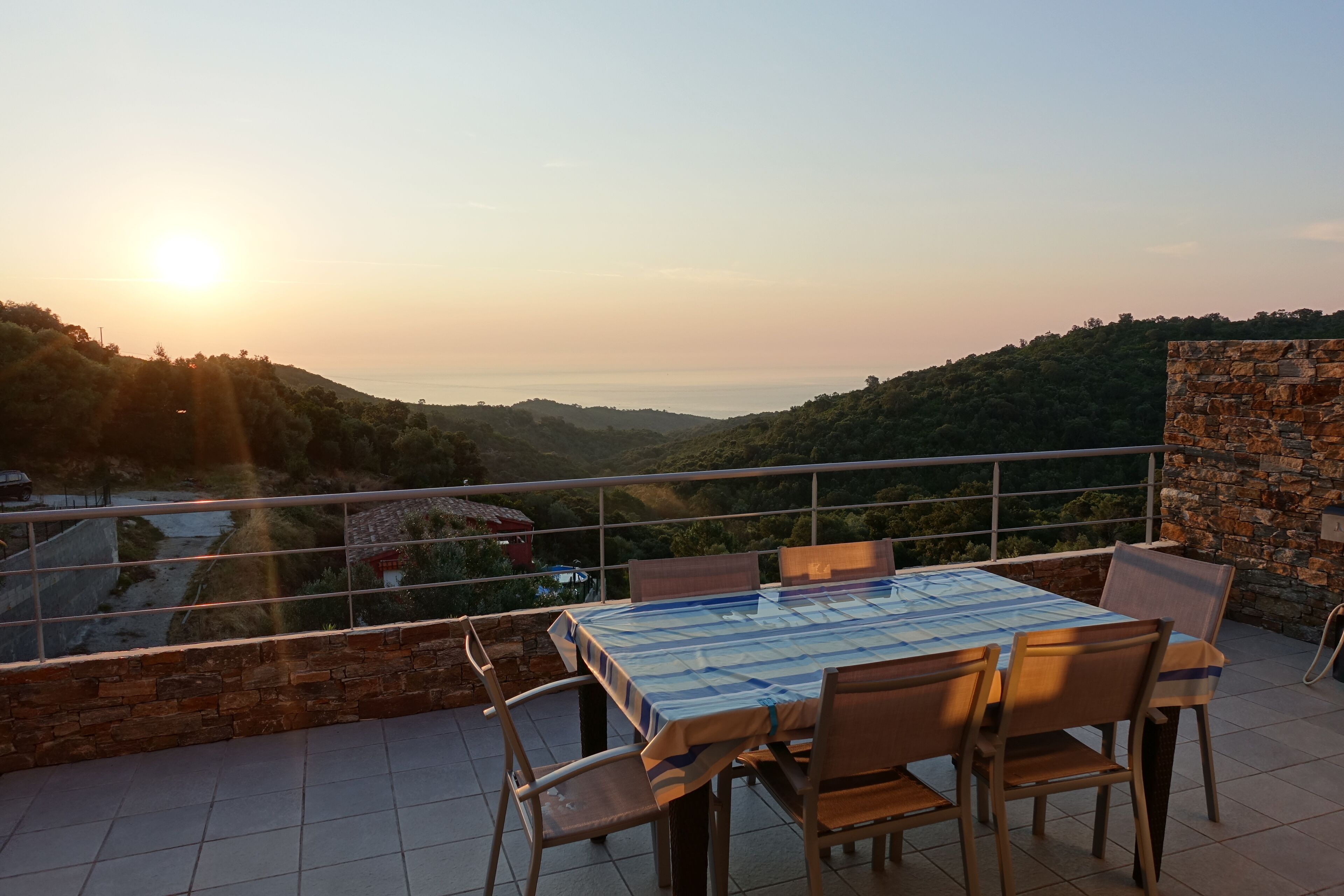 Eine Terrasse mit Tisch und Stühlen, mit Blick auf ein Tal und ein Gewässer bei Sonnenuntergang.
