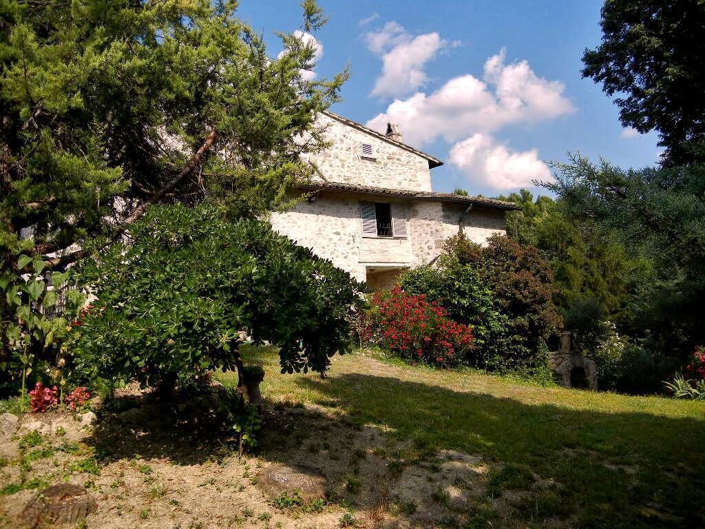 A rustic stone building with a sloped roof, surrounded by lush greenery and flowering plants.