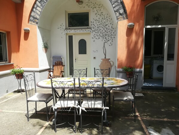 Un espace extérieur aménagé pour les repas, avec une table ronde et des chaises, un pot de fleurs et une porte surmontée d’une fenêtre.