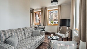 Un salón moderno con un sofá de cuadros, un sillón, una mesa de centro con pasteles, un televisor y una ventana con cortinas.