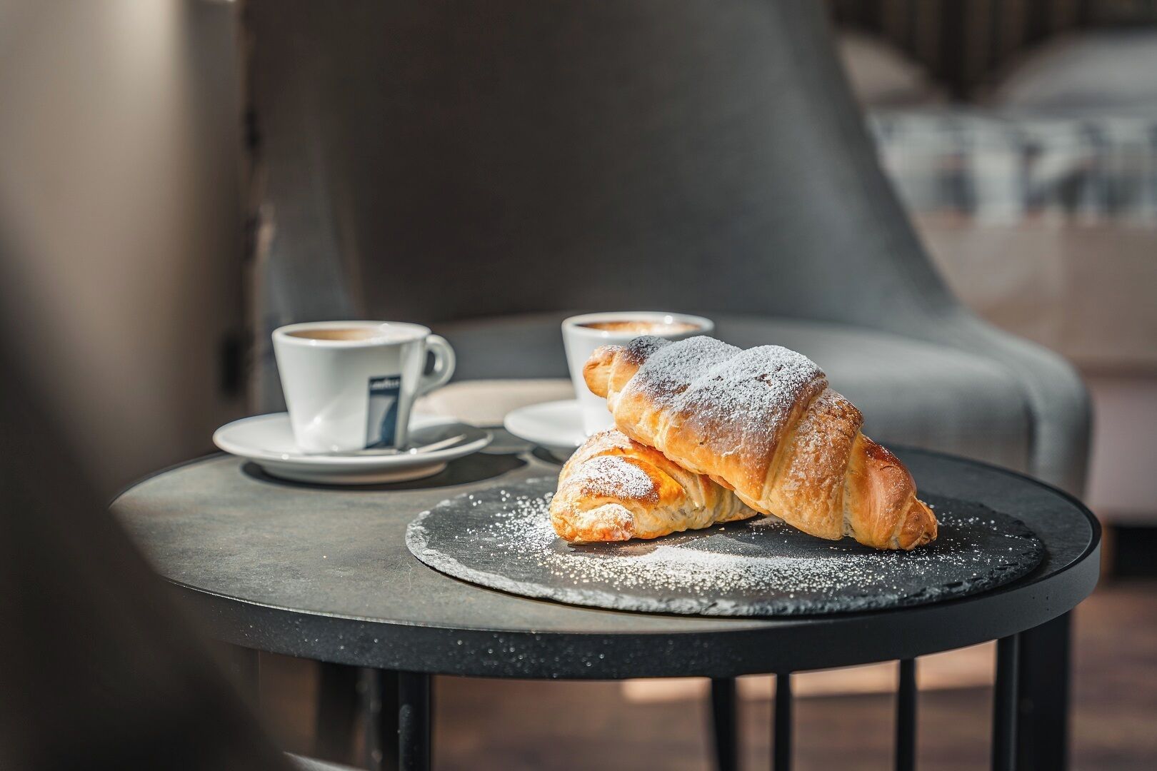A croissant on a plate with powdered sugar, a cup of coffee, and a saucer on a small round table.
