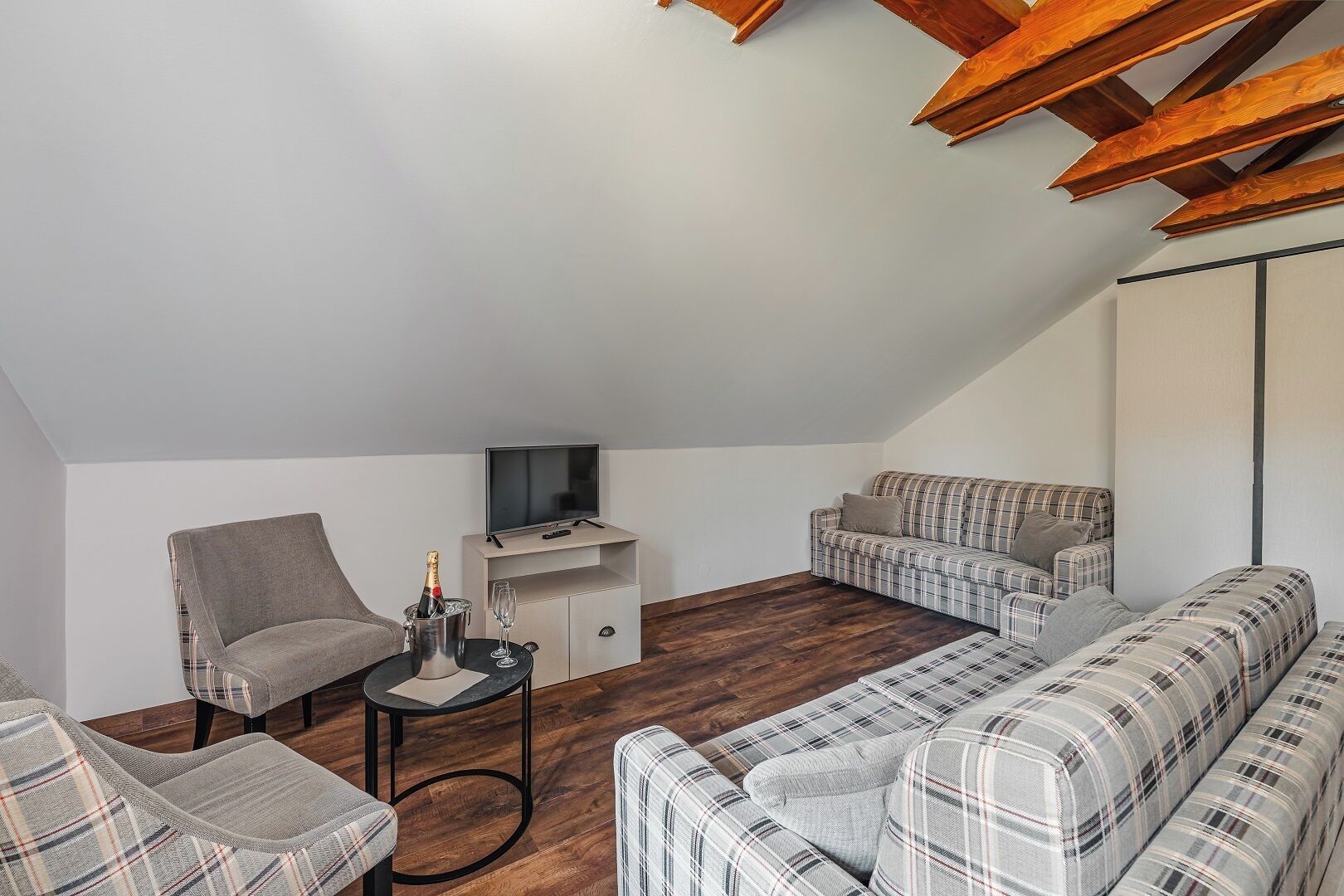 A loft-style living room with a high ceiling, wooden beams, a grey armchair, a plaid sofa, and a TV on a white stand.