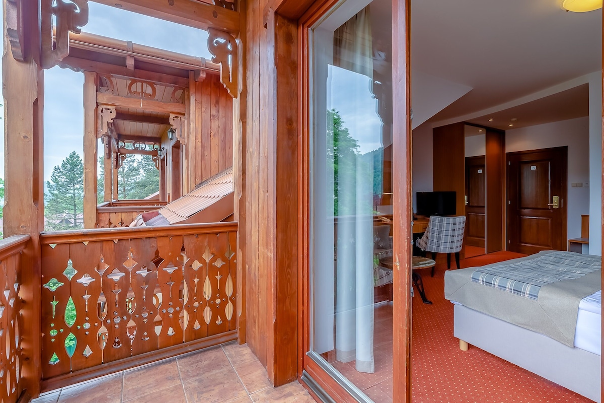 Un balcón de madera con diseños intrincados, un dormitorio con cama y televisor, y un divisor de vidrio corredizo que da al cuarto.