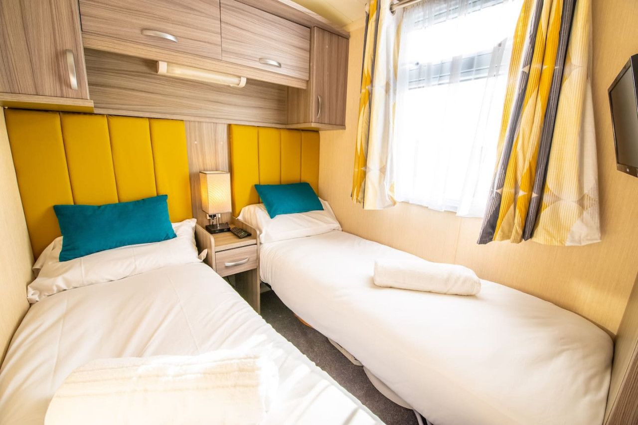 A hotel room with two single beds, a bedside table with a lamp, a TV mounted on the wall, and a window with curtains.