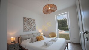 Un dormitorio moderno con una cama grande, lámparas de noche, un elemento decorativo en la pared y vistas a una piscina a través de una puerta abierta.