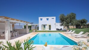 Un área de piscina moderna con piscina azul cristalina, sillones blancos para tomar el sol y una pérgola.