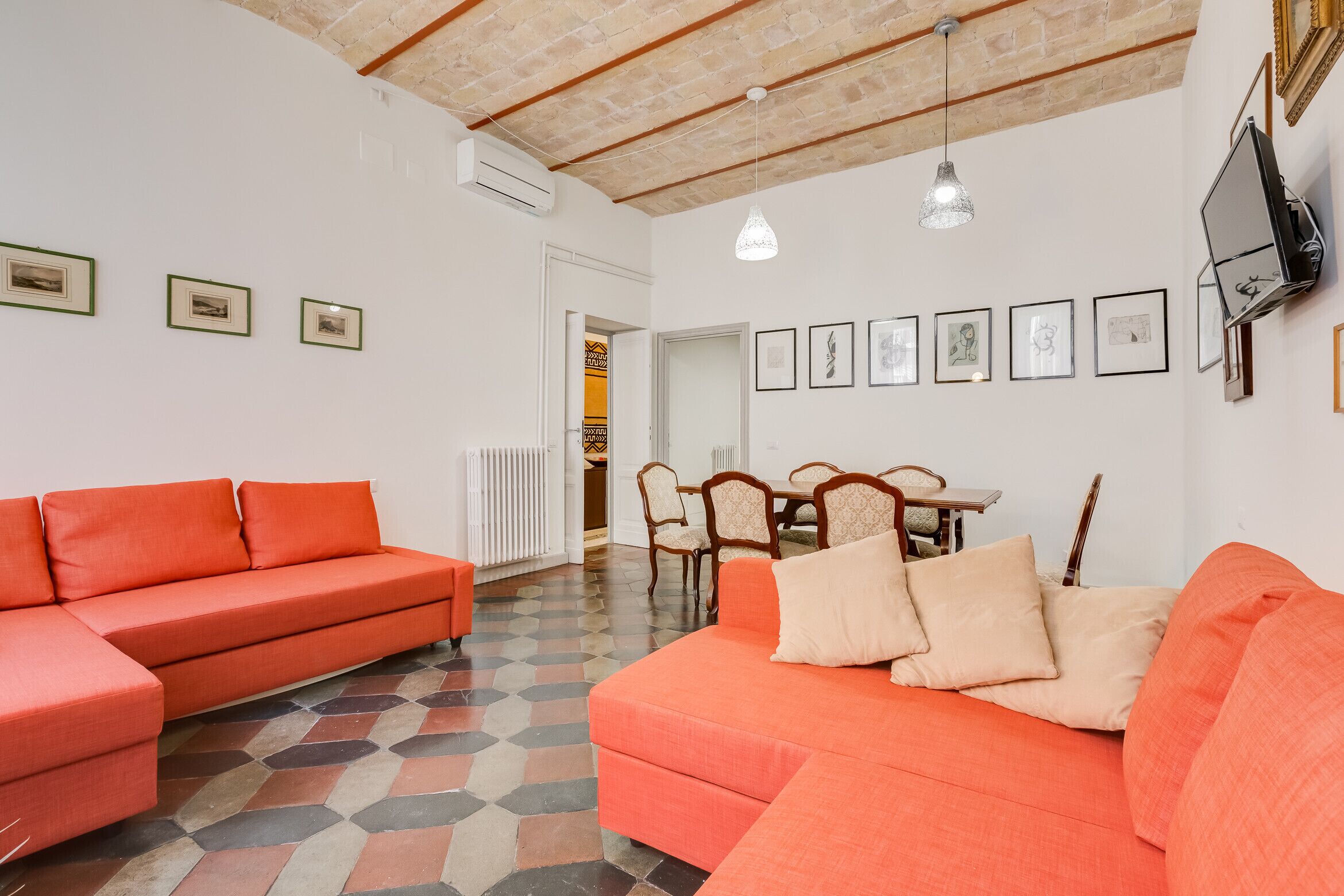 A living room with two orange sofas, a dining table with chairs, and a TV mounted on the wall.