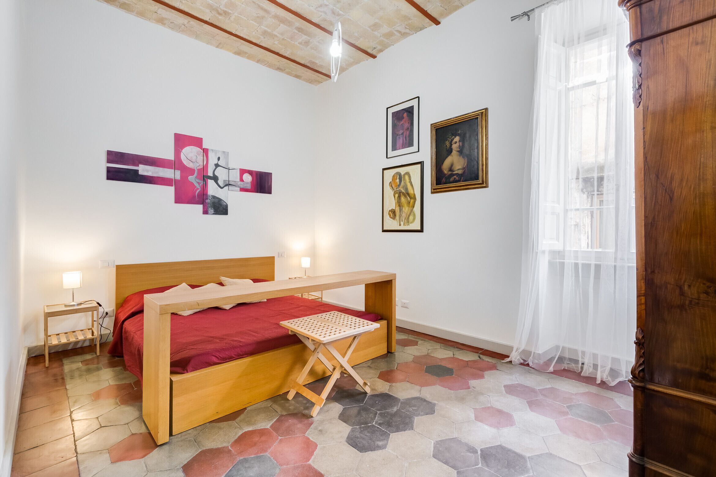 A bedroom with a wooden bed, a small bedside table, a red bedspread, and hexagonal patterned flooring.
