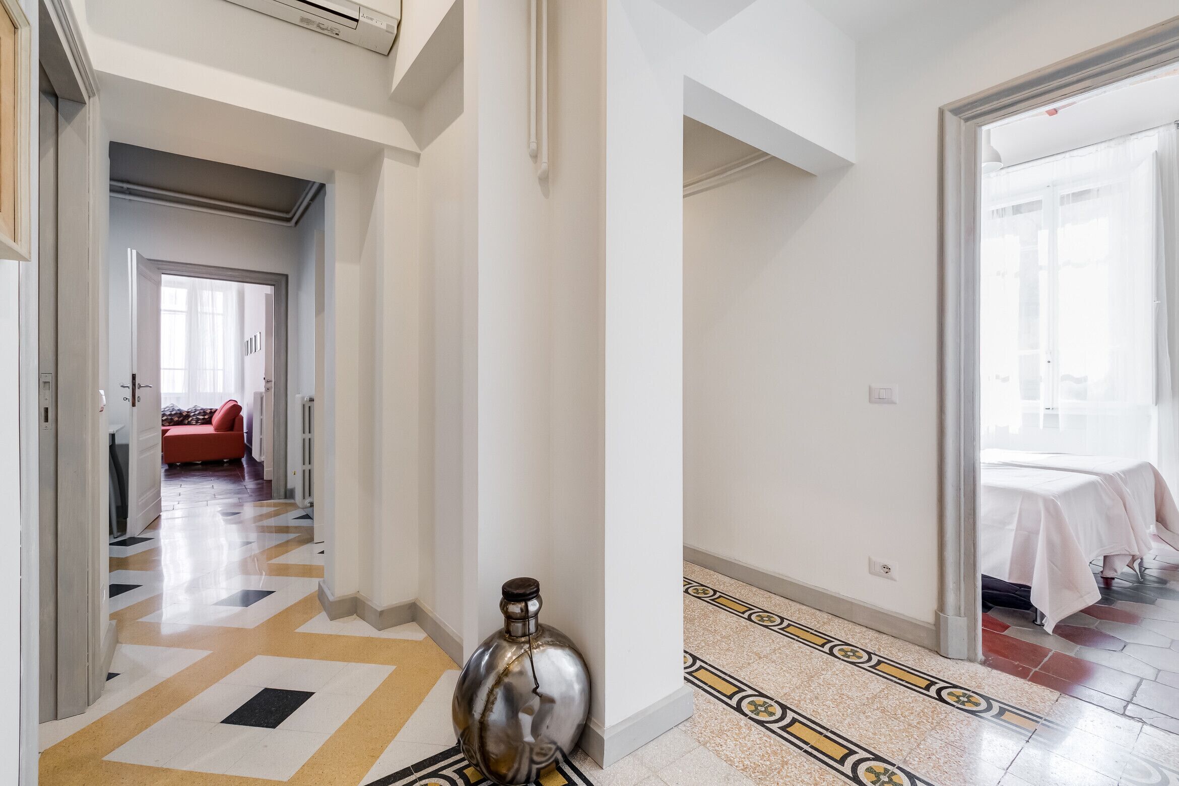 A hotel corridor with a patterned floor, a metallic object, and a bed with a white cover in the adjacent room.