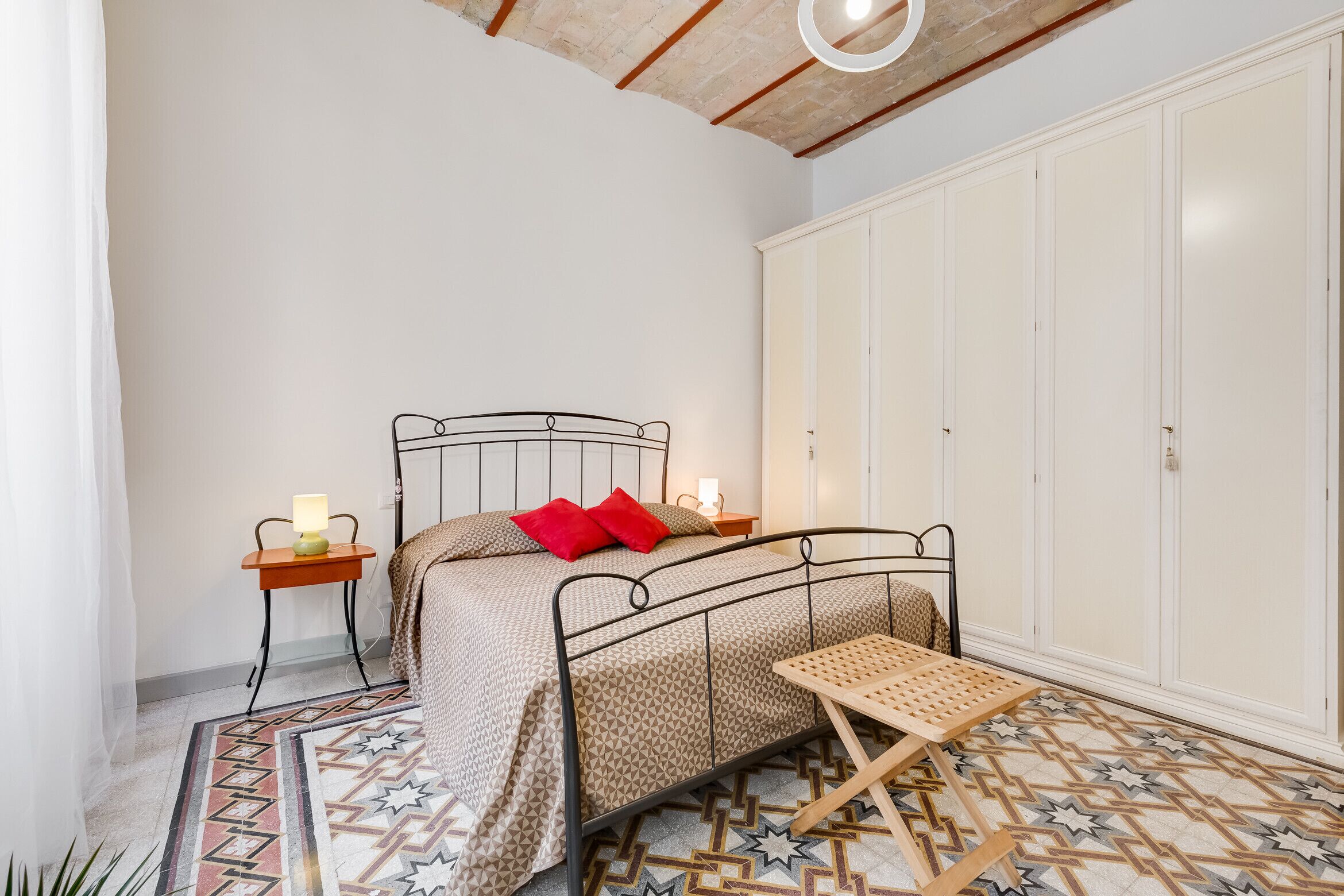 Un dormitorio con estructura de cama metálica, una mesita de noche con lámpara, un armario y un tapete estampado.