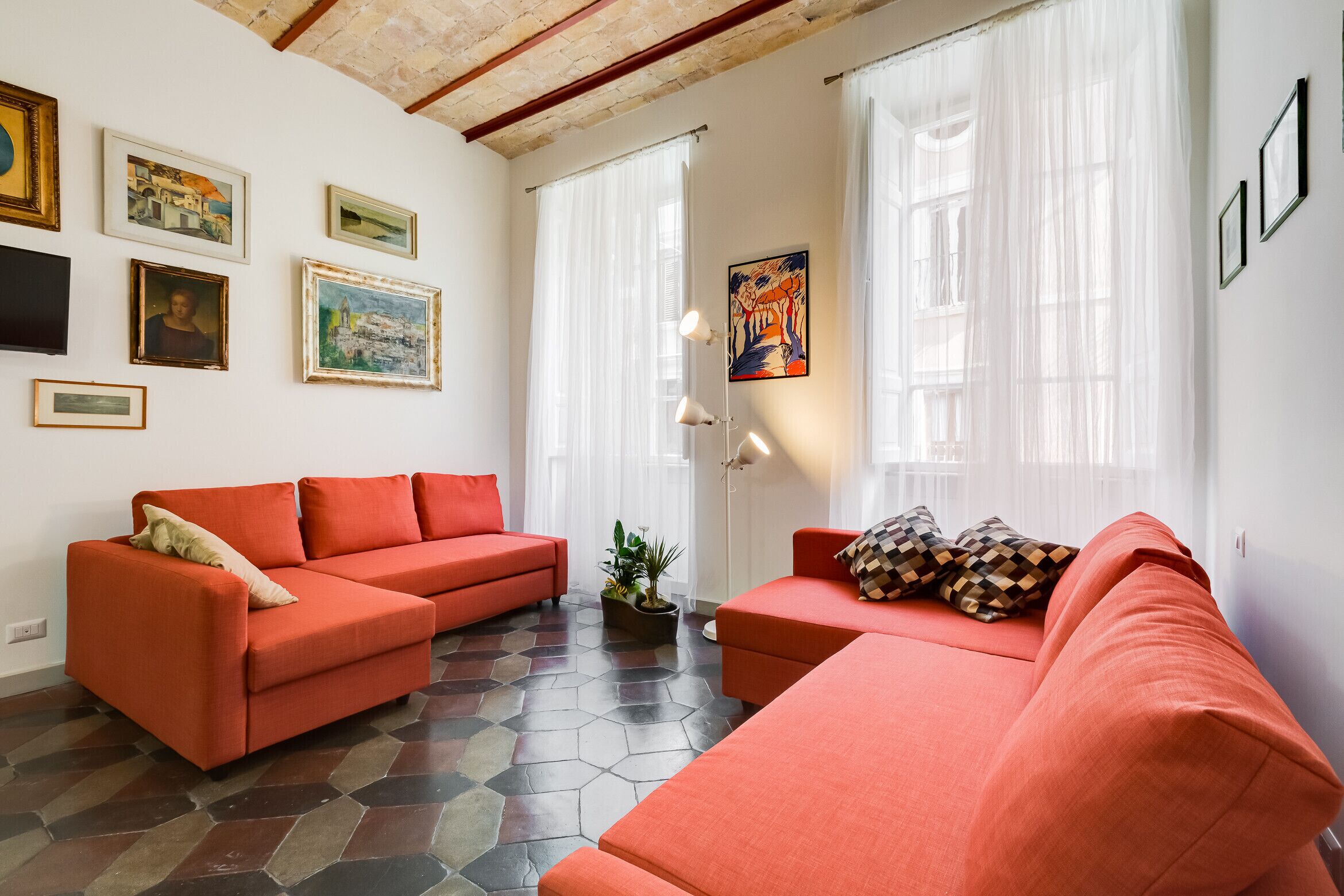 A living room with two red sofas, a coffee table, and a potted plant. There are framed pictures on the walls and a large window with sheer curtains.