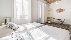 A hotel room with two beds, a small table with chairs, a radiator, and a wall-mounted shelf. - Monti Colosseum Apartment (Roma)