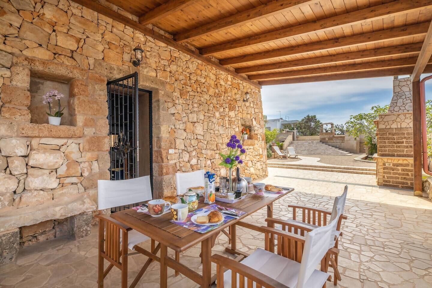 Zona pranzo all'aperto con pareti in pietra, un tavolo e sedie in legno, un camino in pietra e vista su una scala che conduce a un livello superiore.