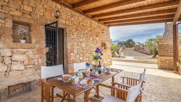Zona pranzo all'aperto con pareti in pietra, un tavolo e sedie in legno, un camino in pietra e vista su una scala che conduce a un livello superiore.