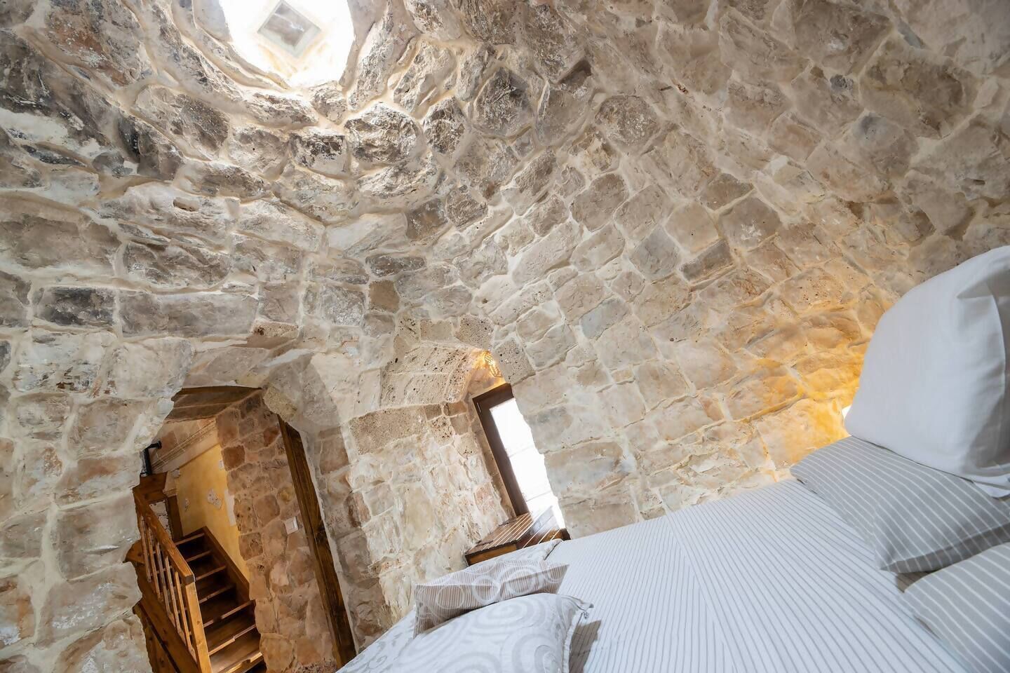A stone-walled room with a bed, a window, and a staircase leading to an upper level.