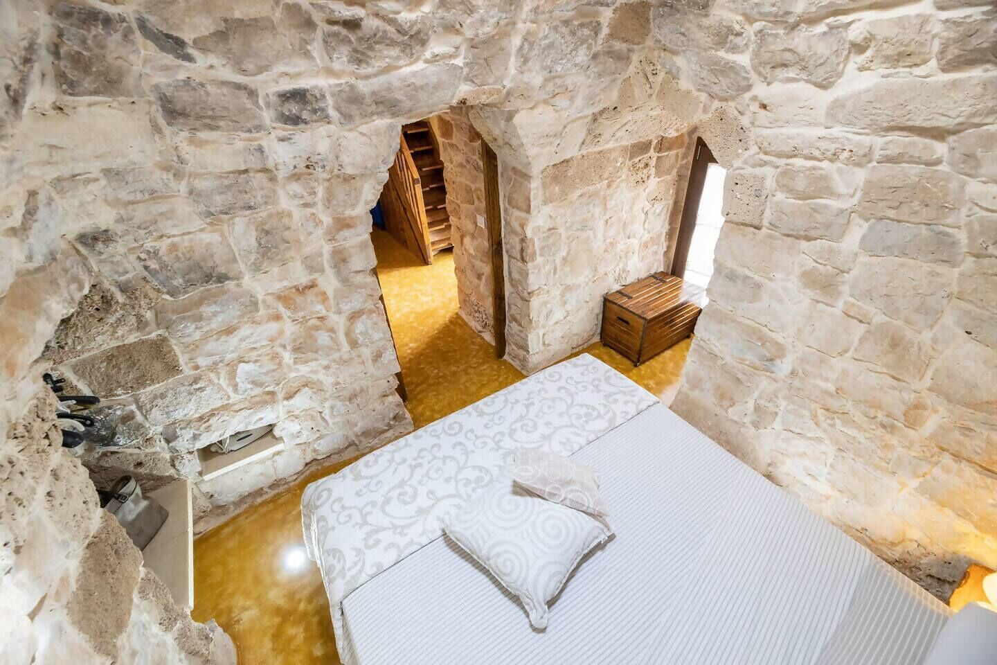 A stone-walled room with a bed, a wooden chest, and a staircase leading to another room.