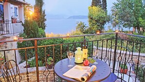 Eine Terrasse mit Tisch für Zwei, komplett mit Weinflasche, Gläsern, Laterne und Speisekarte. Die Terrasse bietet Blick auf ein Gewässer und ist von Grünpflanzen sowie einem Gebäude mit Ziegeldach umgeben.