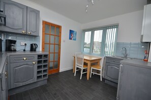 Eine Küche mit grauen Schränken, einer Holztür, einem Esstisch mit Stühlen und einem Fenster mit Jalousien.