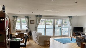A spacious living room with a large window overlooking a harbor, wooden flooring, a sofa, a television, and a dining area with a table and chairs. - 4 bed spacious modern townhouse with spectacular views over the River Tamar.  (Plymouth)
