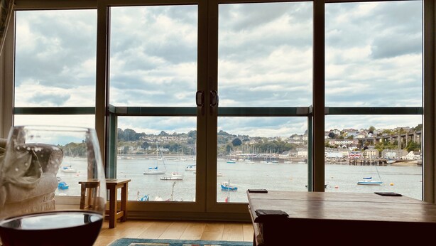 A room with a large window overlooking a harbor, a wooden table, a blue rug, and a glass of red wine. - 4 bed spacious modern townhouse with spectacular views over the River Tamar.  (Plymouth)