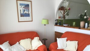 A room with two orange couches, a green floor lamp, a framed picture on the wall, and a kitchen area with a vase of flowers.