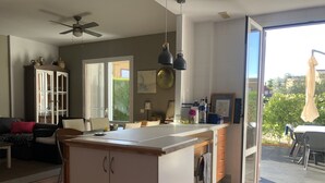 Una cocina moderna con una isla central, un comedor con mesa y sillas, un ventilador de techo y un ventanal corredizo que da a un patio exterior.