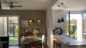 Un comedor con mesa, sillas y un ventilador de techo. Hay dos relojes en la pared y una ventana con vistas al exterior.