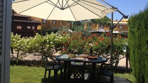 Un patio exterior con mesa y sillas bajo un paraguas grande, rodeado de plantas con flores y un seto cuidado.