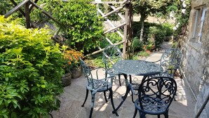 A garden patio with a round metal table and four chairs, surrounded by lush greenery and potted plants.