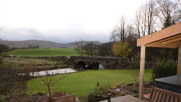 A patio with a table, chairs, and a bottle of wine. A stone bridge and a grassy field with a river are visible in the background. - Spacious Riverside Cottage Perfect For Families, groups,pets. ** NEW HOT TUB** (Glasgow)