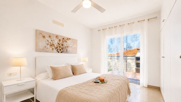 Ein Schlafzimmer mit Bett, Nachttischen, Deckenventilator, einem Gemälde und Blick ins Freie.