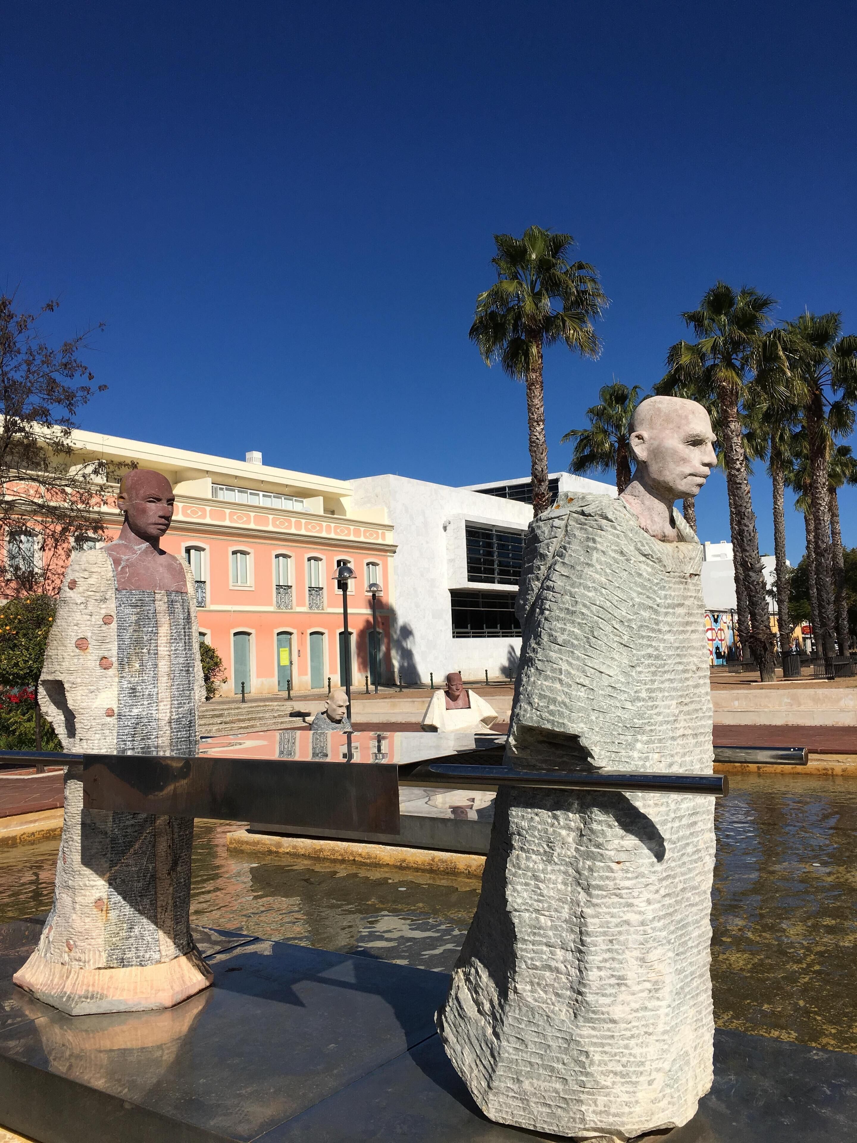 Une sculpture représentant un homme et une femme, placée sur une place publique, avec des palmiers et un bâtiment en arrière-plan.