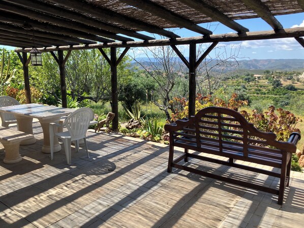 A covered patio with a table and chairs, a wooden bench, and a view of a mountainous landscape.