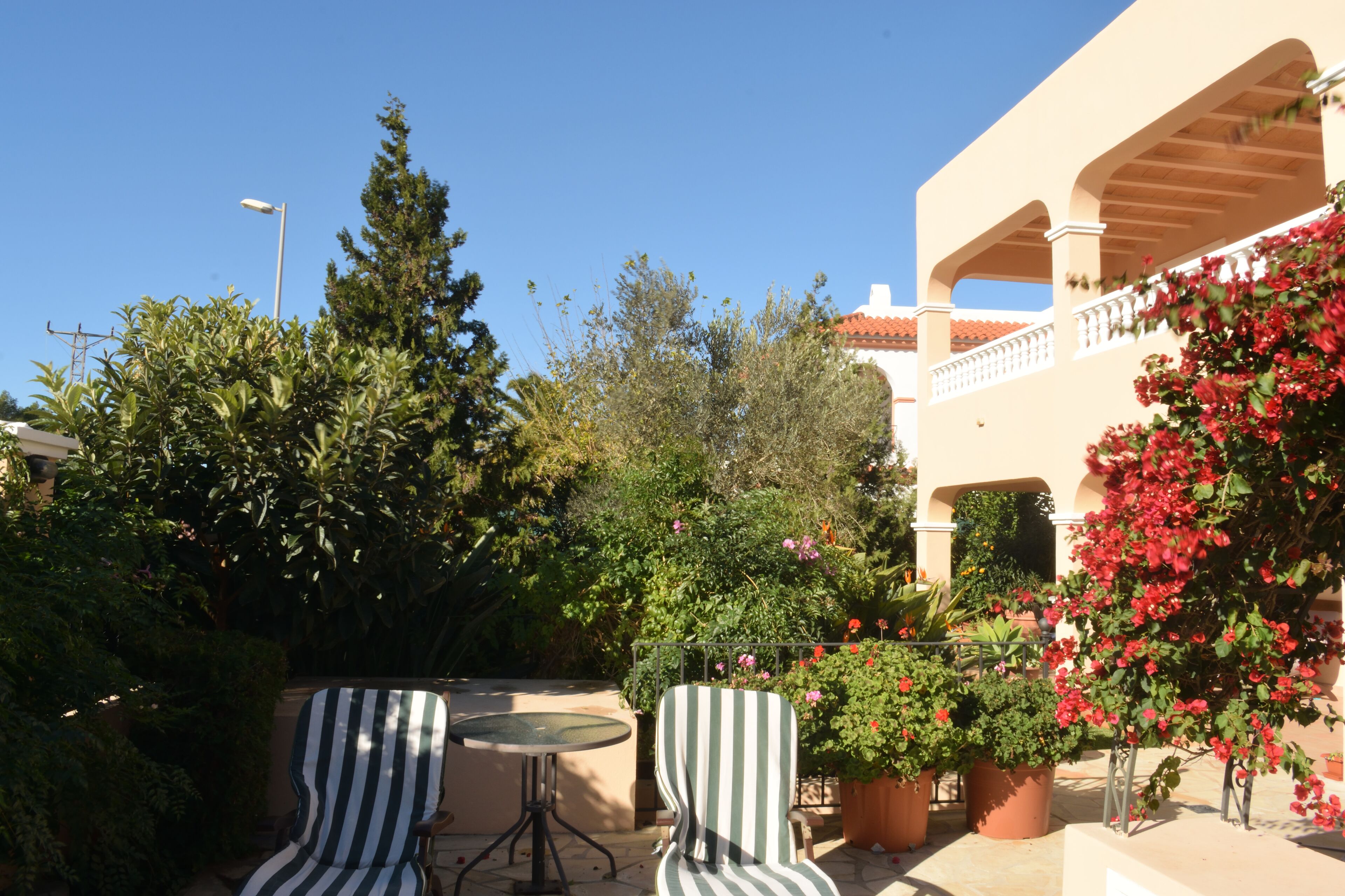 A patio area with striped chairs and a round table, surrounded by potted plants and a building with a balcony.