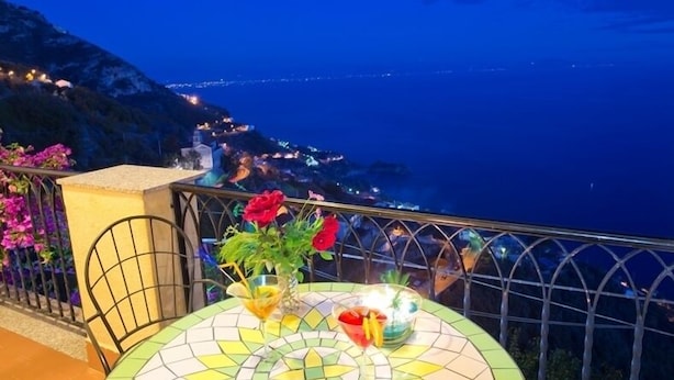 Un balcon avec une table dressée pour deux personnes, donnant sur une ville côtière illuminée la nuit.