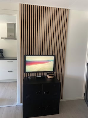 A modern living room with a flat-screen TV on a black cabinet, a wooden slat wall, and a kitchen area with white cabinets and a stainless steel range hood. - Het Paalhoofd 3 personen  (Westkapelle)