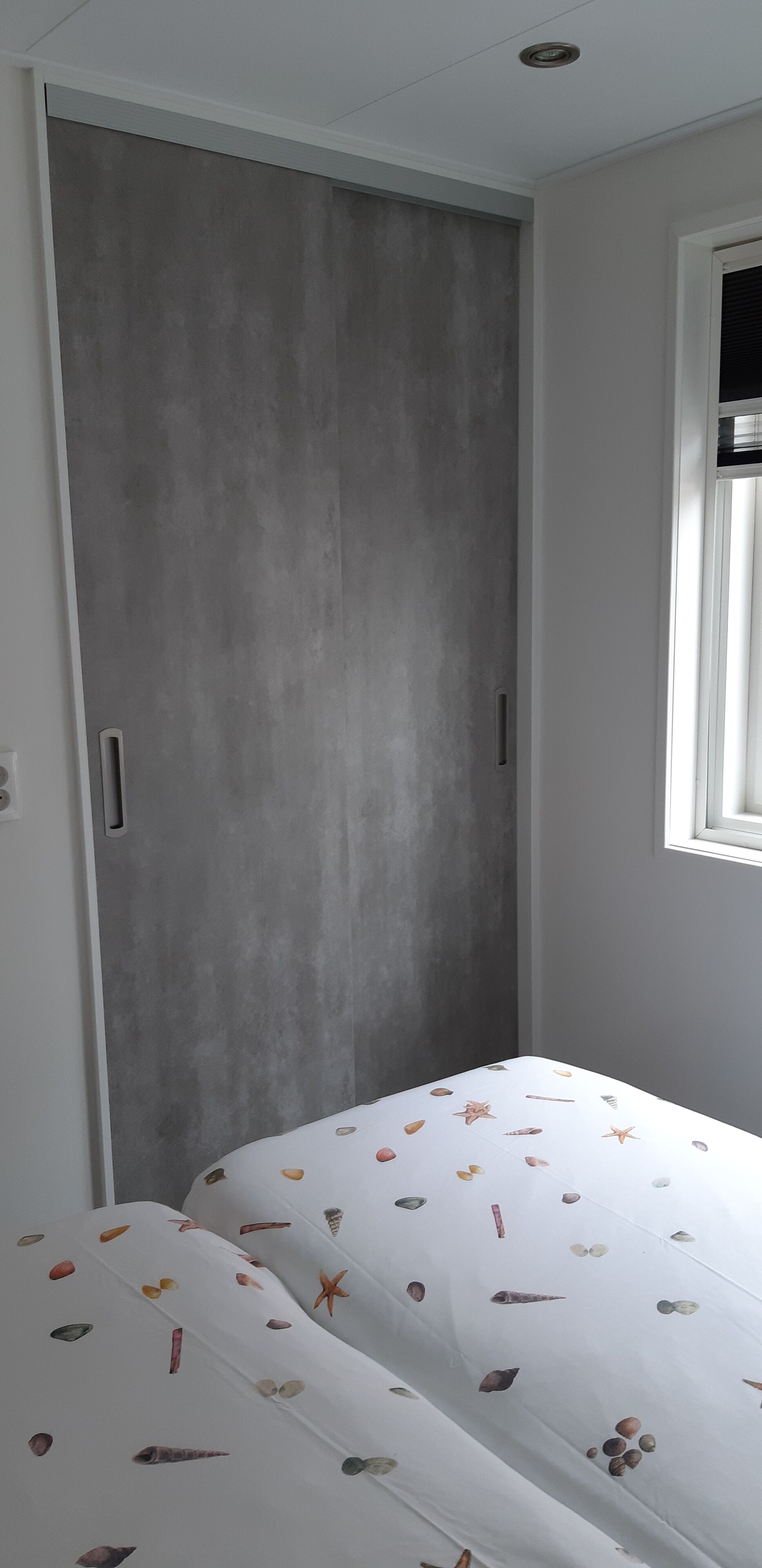 Un dormitorio con un armario gris, una ventana con persianas y una cama con colcha blanca de diseño con motivos de conchas marinas.