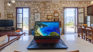 A room with a stone wall, a laptop on a table, a TV, and a dining area with chairs and a table. - Liladi Stone-Built Villa With Exclusive Pool. (Fiskardo)