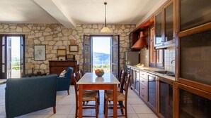 A spacious dining area with a stone wall backdrop, a large window offering a view of the outdoors, and a modern kitchen with wooden cabinets and a hanging light fixture. - Liladi Stone-Built Villa With Exclusive Pool. (Fiskardo)