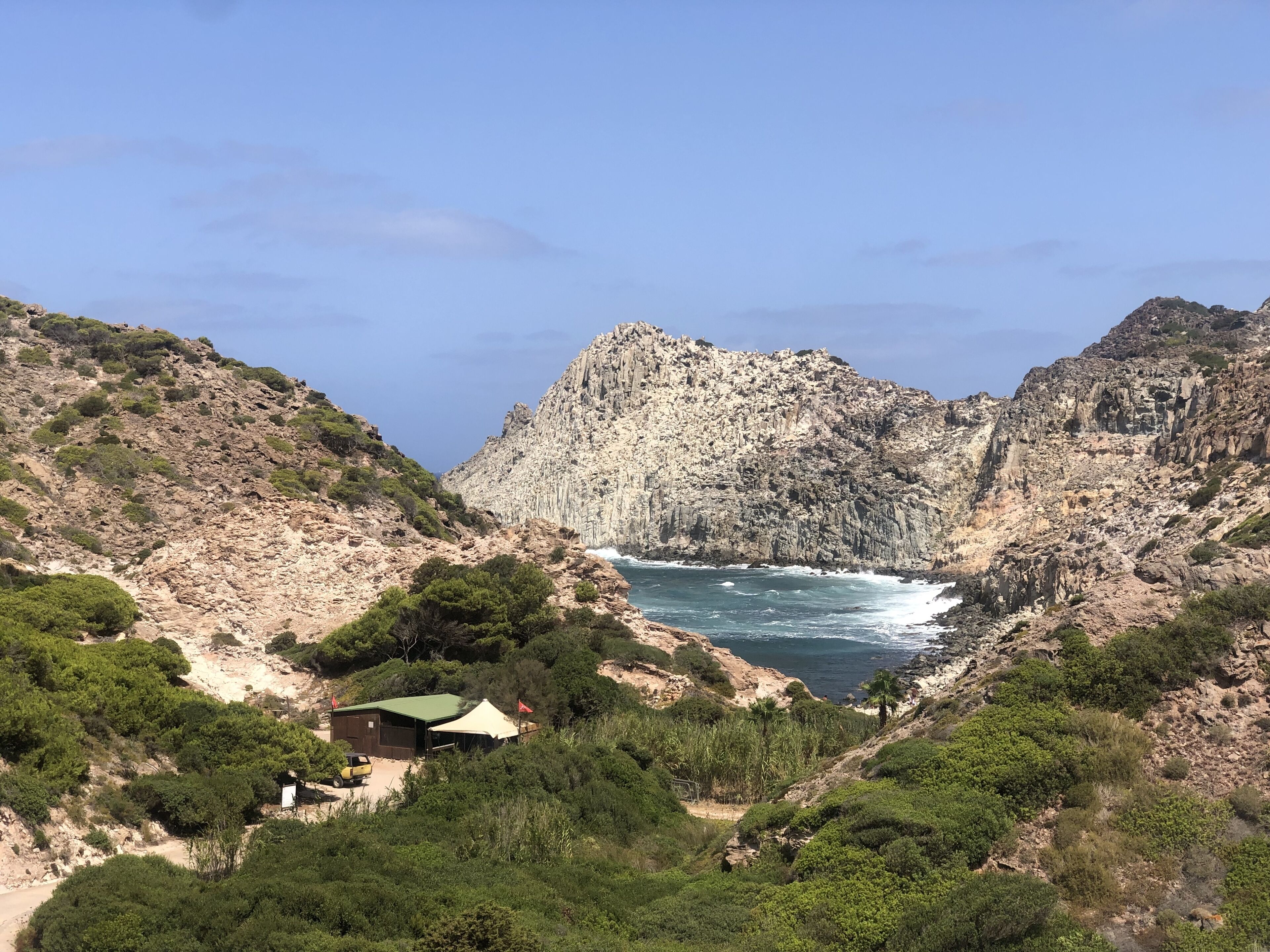 Paisagem costeira acidentada com falésias rochosas, uma praia e um pequeno edifício com telhado verde.