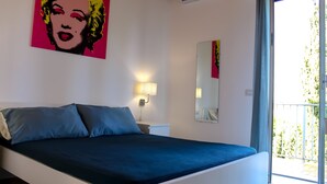 A bedroom with a bed, a wall-mounted lamp, a mirror, and a window with a view of greenery. - Villa Eleonora Home Holiday (Marina di Ragusa)