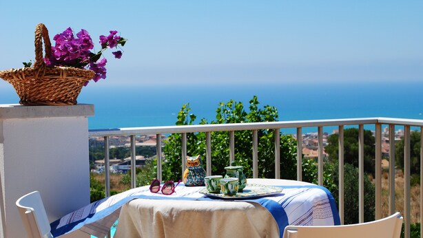 A balcony with a table set for tea, a wicker basket with flowers, and a view of the sea. - Villa Eleonora Home Holiday (Marina di Ragusa)
