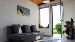 A modern living room with a grey sofa, a patterned rug, a painting on the wall, and a view of greenery outside. - Villa Eleonora Home Holiday (Marina di Ragusa)
