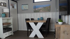 Uma área de refeições com mesa de madeira e cadeiras pretas, base branca em formato de X, armário branco com gavetas e prateleira com cestos.