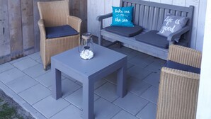 Uma cabana de madeira com varanda, com mobiliário de vime, uma mesa de vidro e um pote, e vista para a praia através de uma janela.