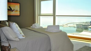 A hotel room with a large window offering a view of a marina and boats. The bed is neatly made with a grey polka dot bedspread and white pillows.