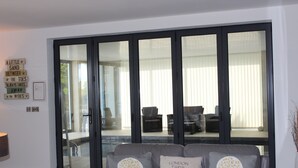 A modern living room with a grey sofa, two decorative pillows, a wall-mounted lamp, and a large sliding glass door leading to an outdoor seating area with a pool. - Luxury Apartment - Sleeps 4 -  Spectacular Views with Private Pool and Hot Tub   (Torquay)