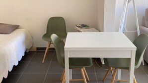A room with a white bed, a framed black and white photograph on the wall, a white table with green chairs, and a floor lamp. - Luxury Apartment (Collioure)