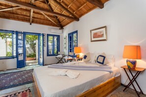 Ein Schlafzimmer mit Holzboden, einem Bett mit weißer Bettwäsche, zwei Nachttischlampen, einem gerahmten Bild an der Wand und Blick ins Freie durch offene Türen.